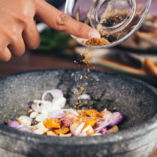 Hand sprinkling spices over a bowl of chopped vegetables on a wooden surface