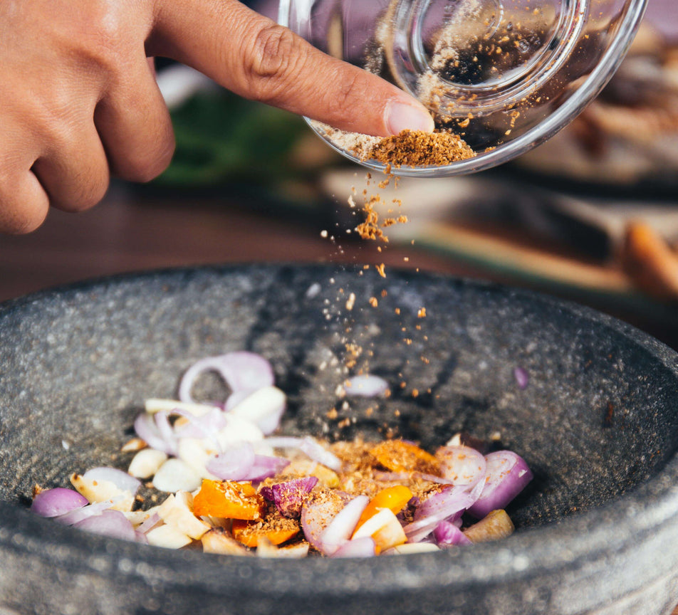 Hand sprinkling spices over a bowl of chopped vegetables on a wooden surface